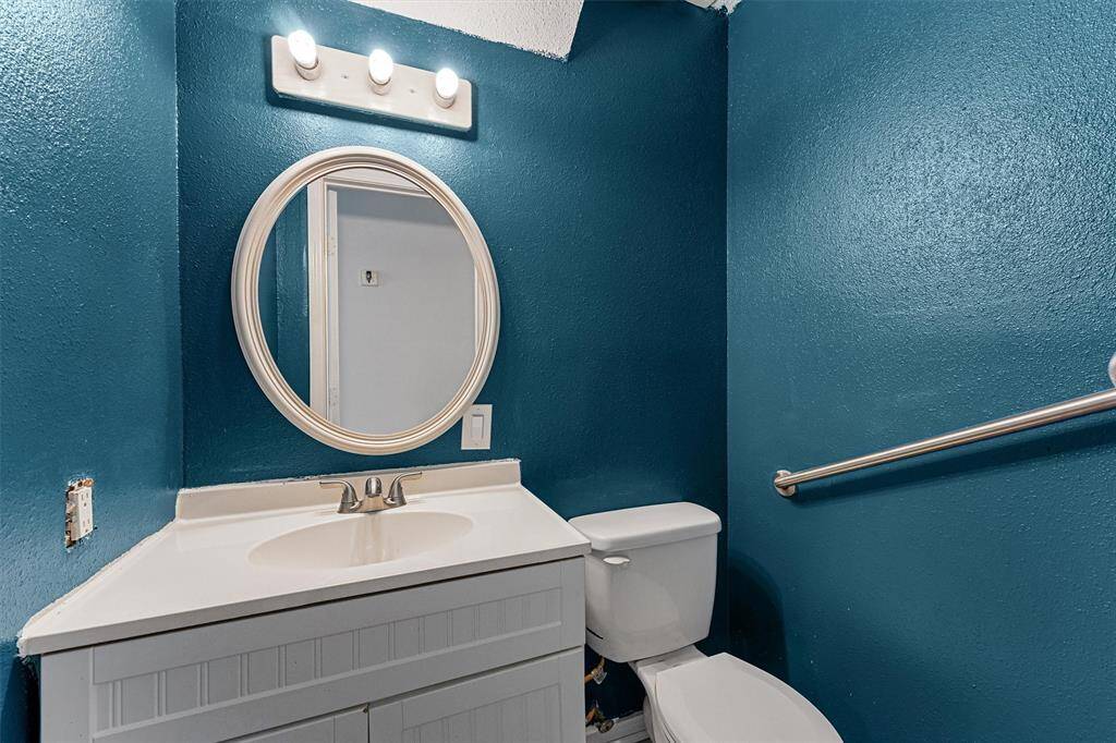 A compact bathroom with rich teal walls, featuring a white vanity and sink, a round mirror, and a toilet. The space is accented by a simple light fixture and a metal grab bar.
