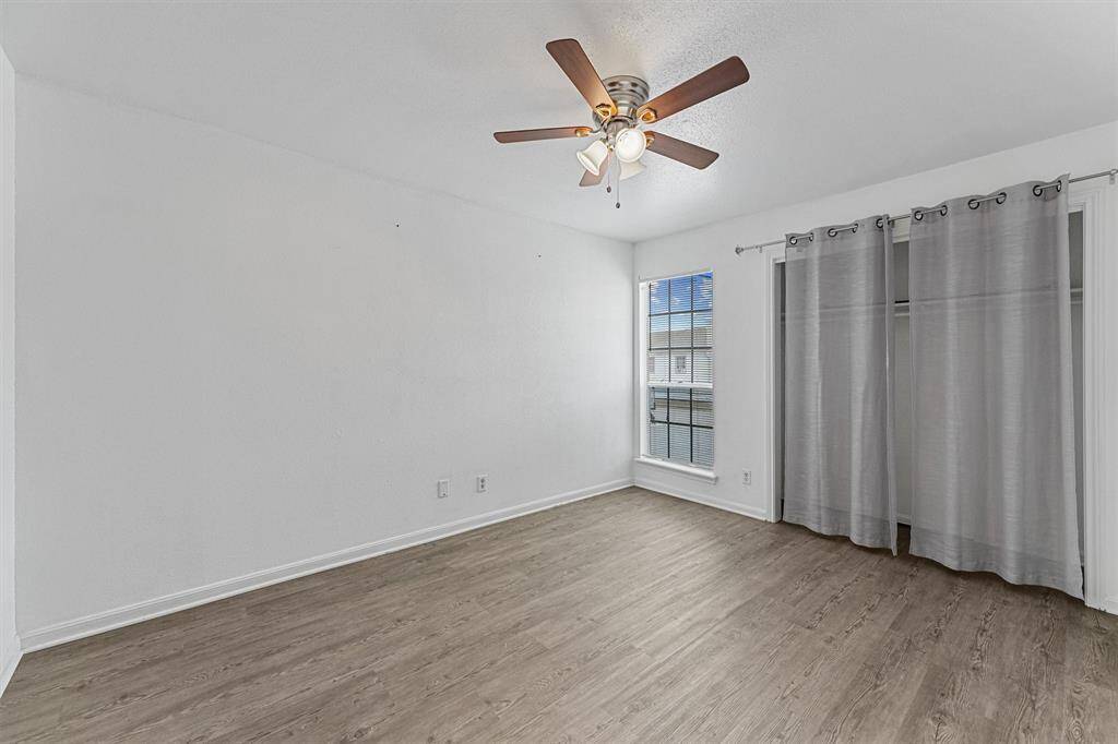 This room features a light, modern design with wood-look flooring, a ceiling fan, and ample natural light from a large window. It includes a spacious closet with curtain doors.