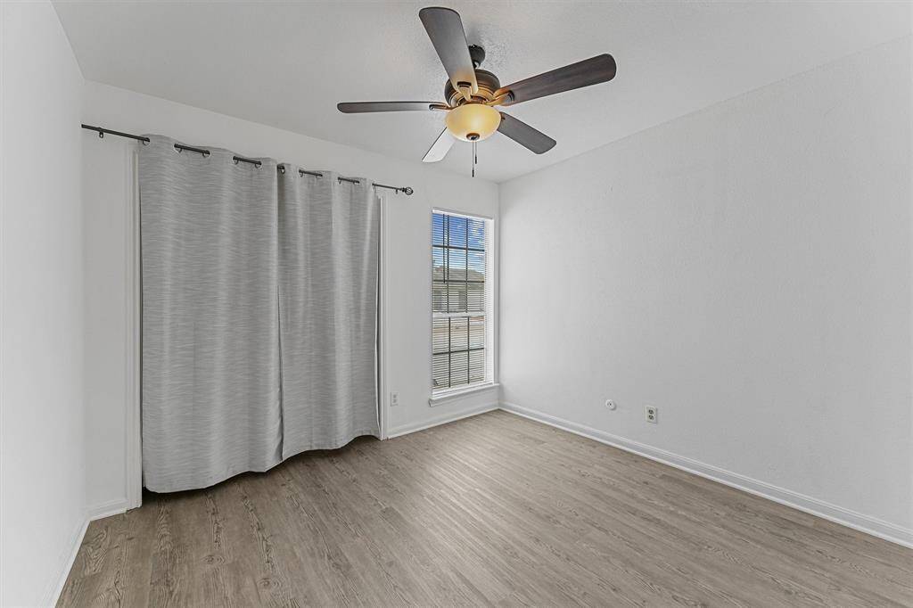 This room features a modern ceiling fan, light wood flooring, and a large window with natural light and blinds. The walls are painted white, and there's a curtain for added privacy.