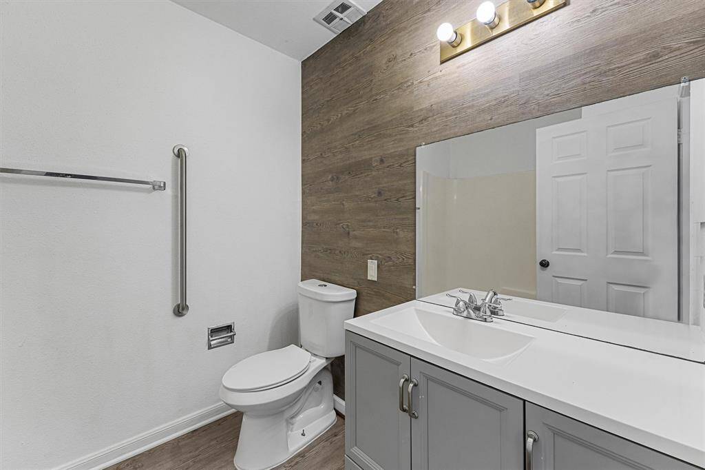 This bathroom features a modern design with a wood-accented wall, large mirror, sleek vanity with dual faucets, and a standard toilet. The lighting is contemporary, complementing the neutral color scheme.