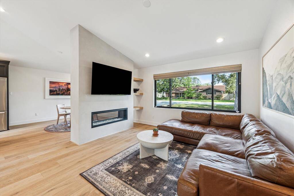 The living room features a vaulted ceiling and a wall of windows that flood the space with natural light. White oak shelving separates the room from the dining area and is anchored to the limewashed double-sided gas fireplace.