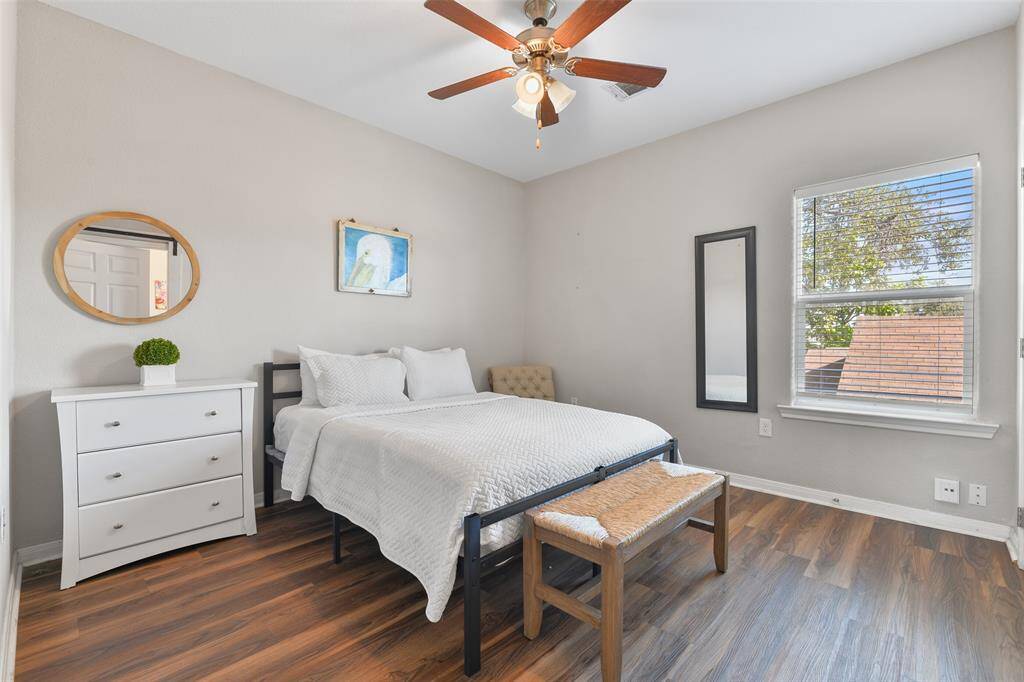 This master suite features a cozy bed with a white duvet, complemented by a luxury vinyl plank floor and light neutral wall paint. It includes a ceiling fan, a small dresser with a round mirror, and a window providing natural light.
