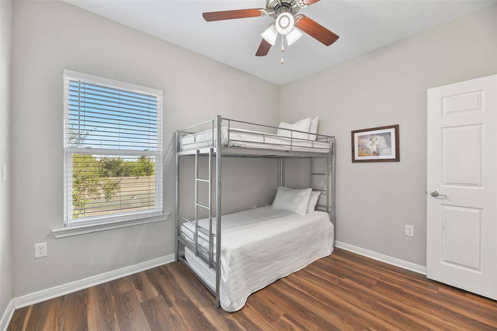 This sunlit bedroom features a bunk bed with a sleek metal frame, a ceiling fan, and easy maintenance luxury vinyl plank flooring. A large window provides natural light and a pleasant view, creating a welcoming atmosphere.