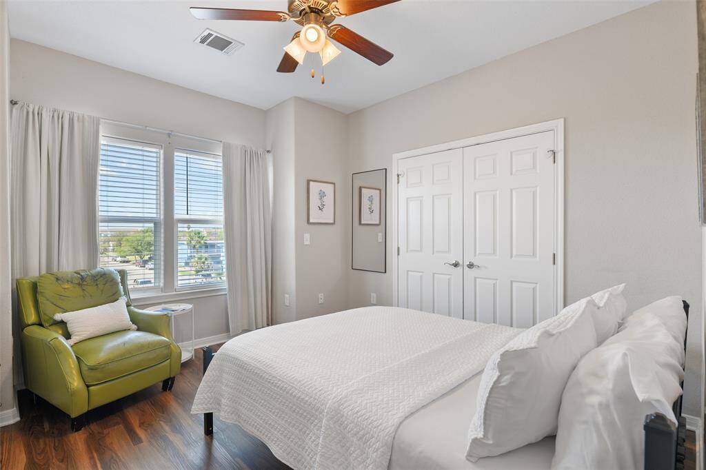 The fourth and final bedroom offers a large window with natural light, neutral wall paint, a comfortable full size bed, and a cozy green armchair. The room includes a ceiling fan and ample closet space.