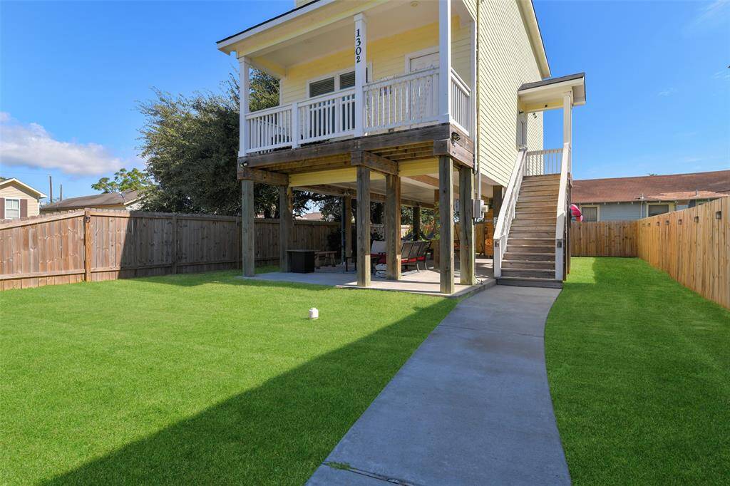 This raised two-story home boasts a spacious, fenced backyard as well as a balcony, an external staircase, and a covered patio area underneath. The yard has a concrete pathway and is well-maintained with lush grass.