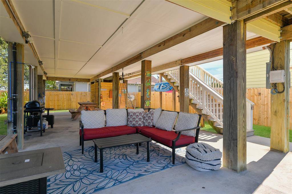 This photo showcases a spacious covered patio with a cozy seating area and rug, a barbecue grill, picnic table, an enclosed outdoor shower, and wooden stairs leading to the living space. The area is fenced for privacy, making it perfect for entertaining.