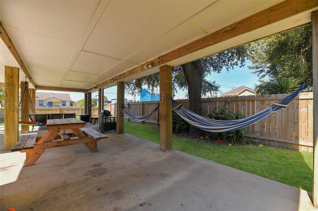 This photo highlights the covered outdoor entertaining space with a concrete floor, a picnic table, hanging swing, grill, dartboard, outdoor shower. Also note the included hammocks & kayak!