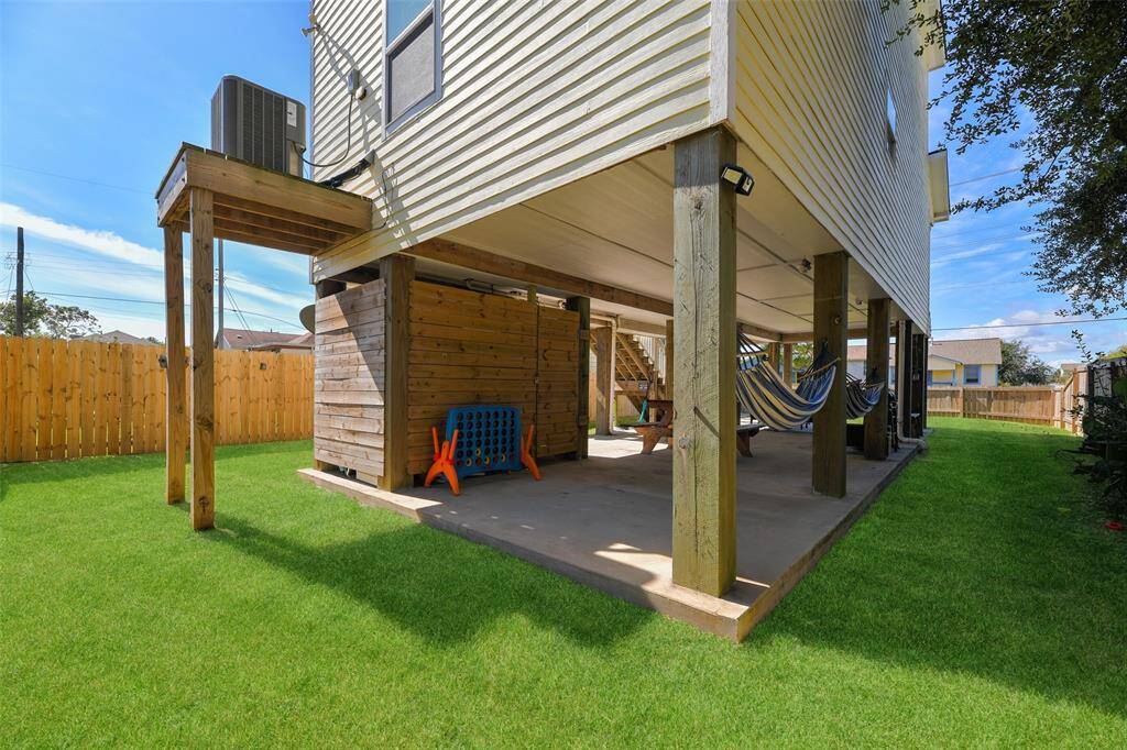 This raised two-story home boasts a spacious, fenced backyard as well as a balcony, an external staircase, and a covered patio area underneath. The yard has a concrete pathway and is well-maintained with lush grass.