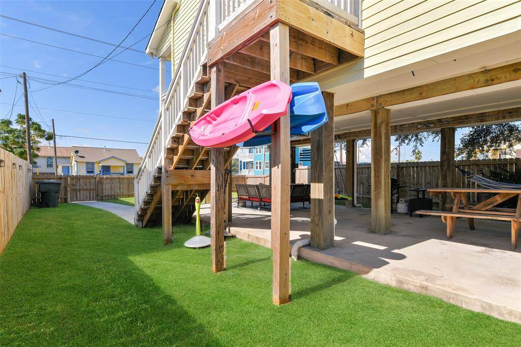 This raised two-story home boasts a spacious, fenced backyard as well as a balcony, an external staircase, and a covered patio area underneath. The yard has a concrete pathway and is well-maintained with lush grass.