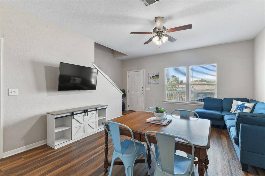 Enter the home into a bright, open living area with wood-look luxury vinyl plank flooring and outfitted with a cozy sectional sofa, a mounted TV, and a dining table. Large windows provide plenty of natural light, and a ceiling fan adds comfort.