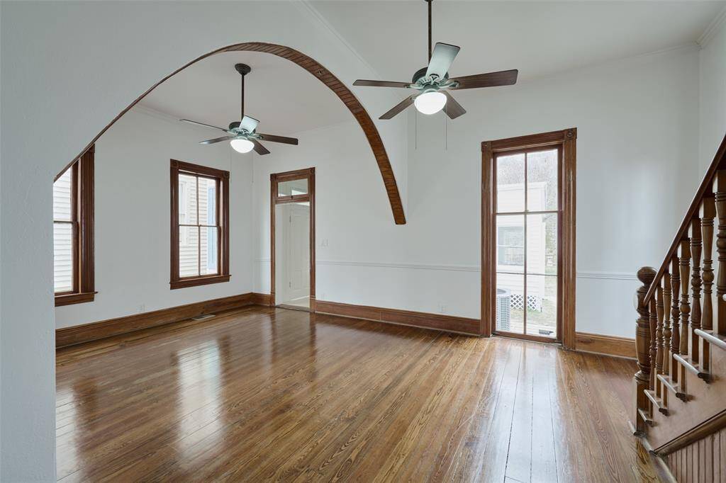 This photo showcases a bright, open space with polished hardwood floors, wooden trim, and ceiling fans. The area features a charming wooden staircase and large windows that provide ample natural light. The adjacent living room with a decorative window pattern adds elegance and character.