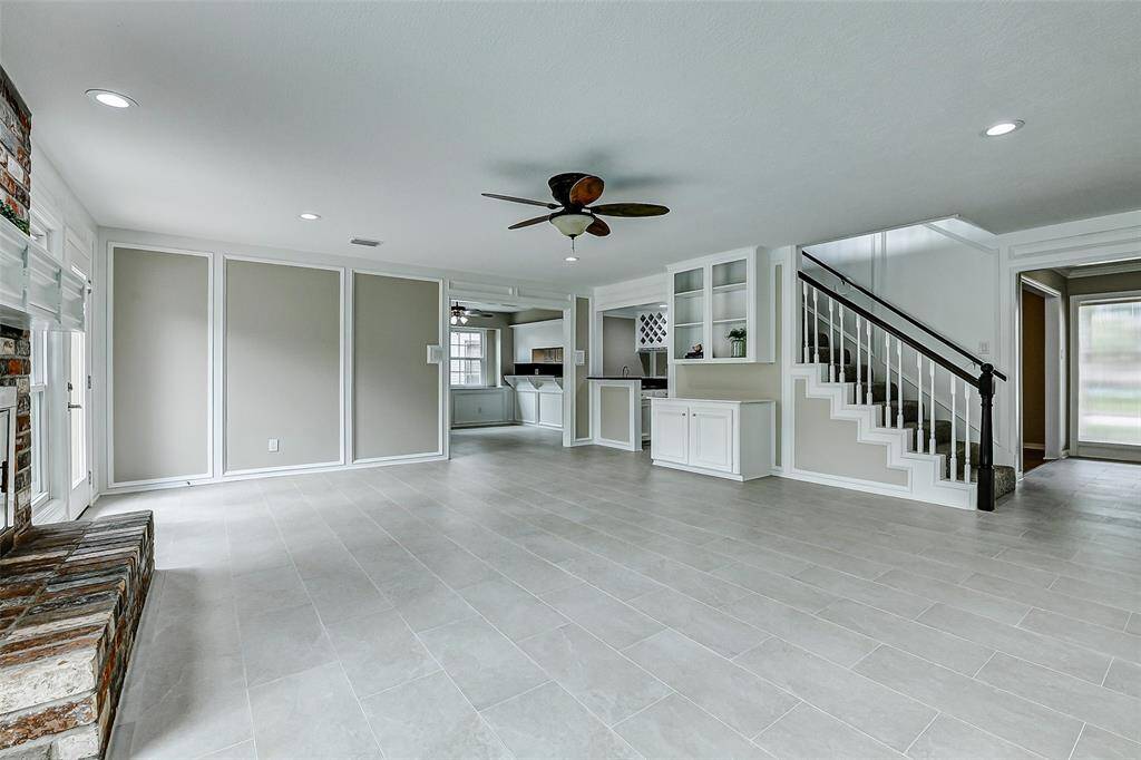 This spacious living area features modern tile flooring, a ceiling fan, and built-in shelving. It includes a stairway with elegant railing, and an open view into the kitchen space, creating a welcoming and functional layout.