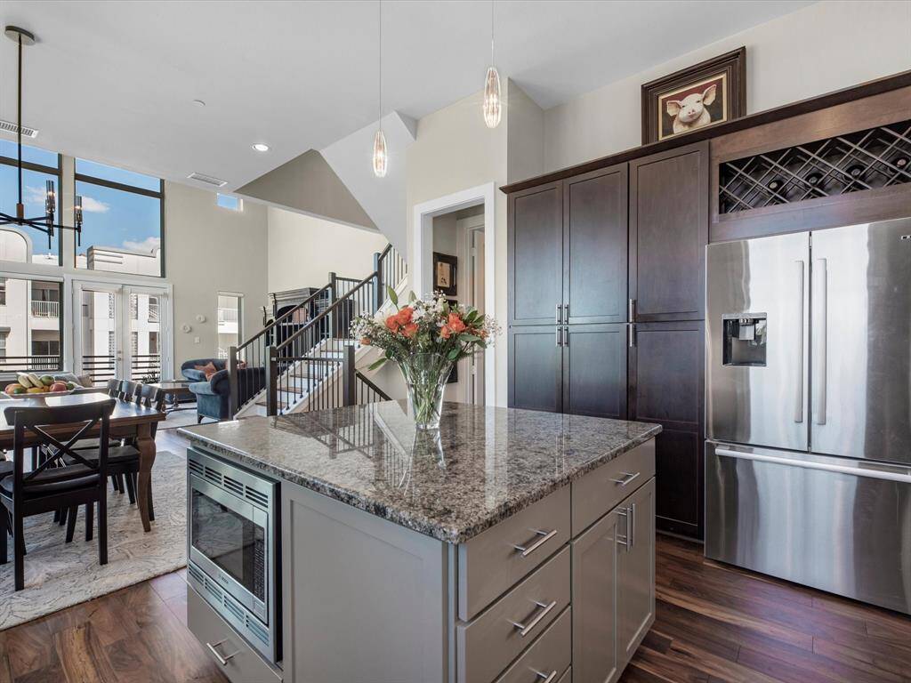 This modern kitchen features a spacious open layout with a large granite island, stainless steel appliances, and dark wood cabinetry. It opens into a bright dining and living area with high ceilings and large windows, offering plenty of natural light.