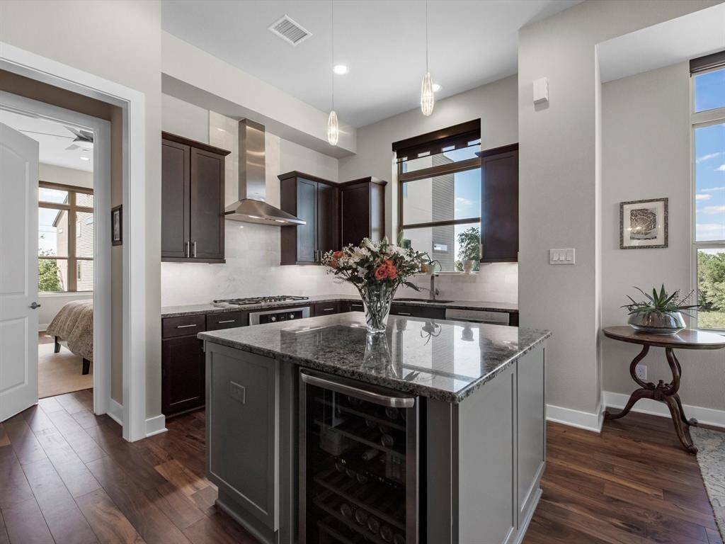 This modern kitchen stands out with dark custom cabinetry, a granite island featuring a built-in wine cooler, and premium stainless appliances. The open layout flows easily into the living space, with natural light and hardwood floors adding warmth and style throughout.