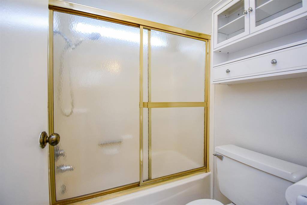 Bathroom features a shower-tub combo with gold-framed sliding glass doors, a white toilet, and wall-mounted storage with glass-paneled cabinets, offering a clean and functional space.