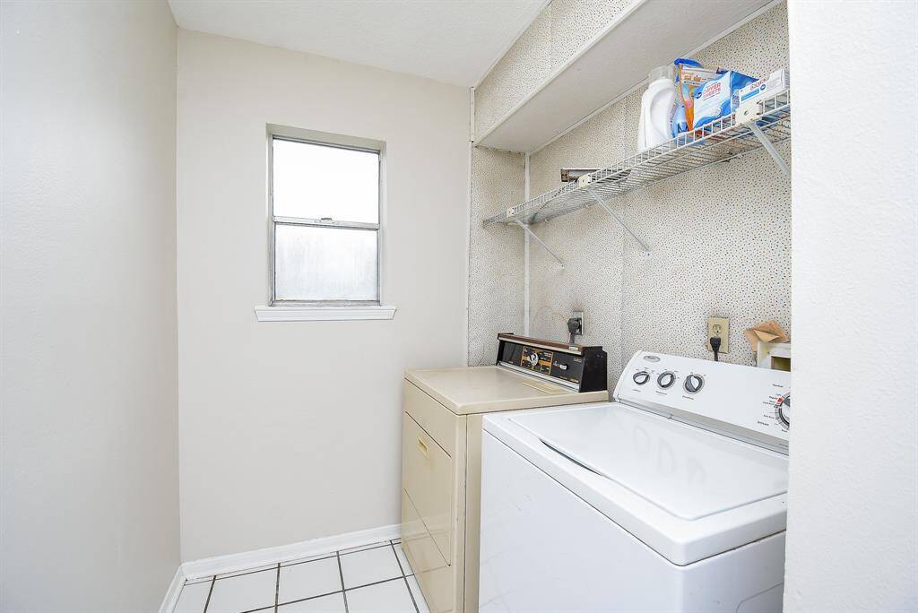 Compact laundry room with washer and dryer, featuring a window for natural light. Includes wall shelves for storage and tiled flooring.