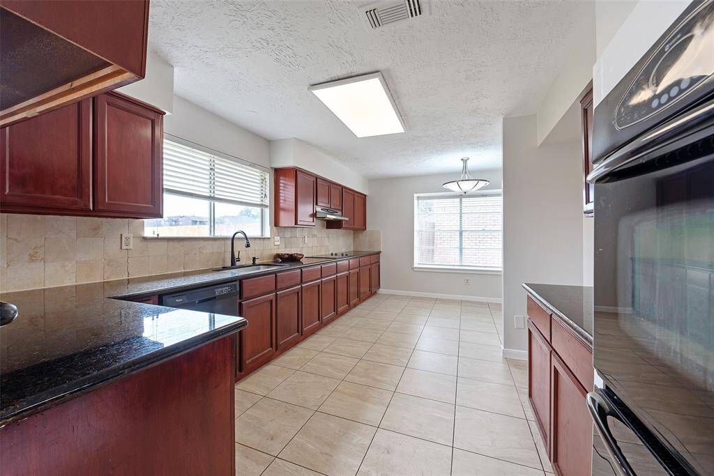 This spacious kitchen features dark wood cabinets, granite countertops, and modern appliances. It has ample natural light from large windows and an adjacent dining area with tile flooring.