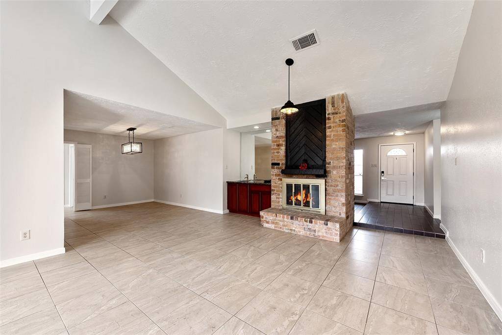 This photo shows a spacious living area with high ceilings and a central brick fireplace. The room features large floor tiles, and there's a dining area to the left with a modern light fixture.