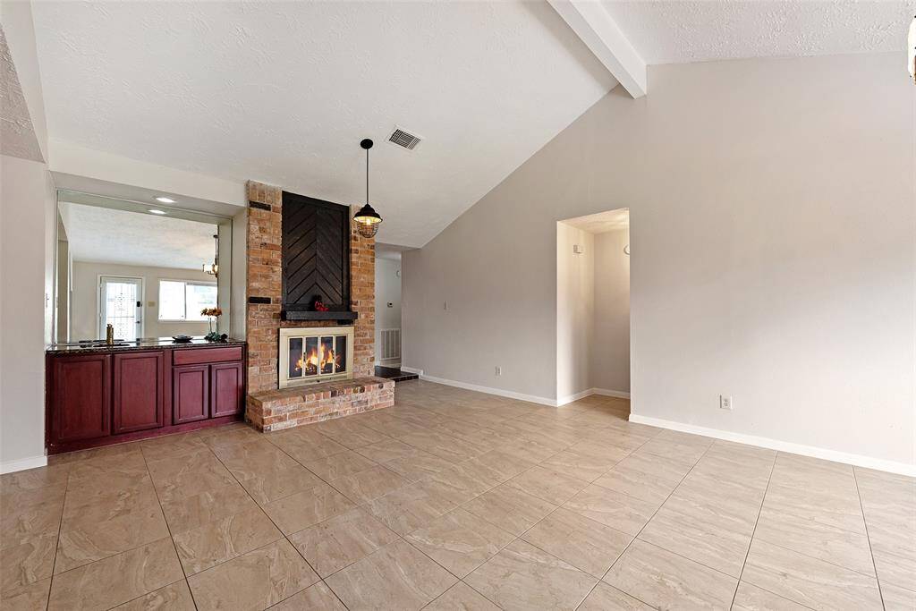 This spacious living room features a vaulted ceiling, a cozy brick fireplace, and large tiled floors. It includes a built-in mirrored bar area with cabinetry, adding a touch of elegance and functionality. The neutral walls offer a versatile backdrop, perfect for personalizing the space.