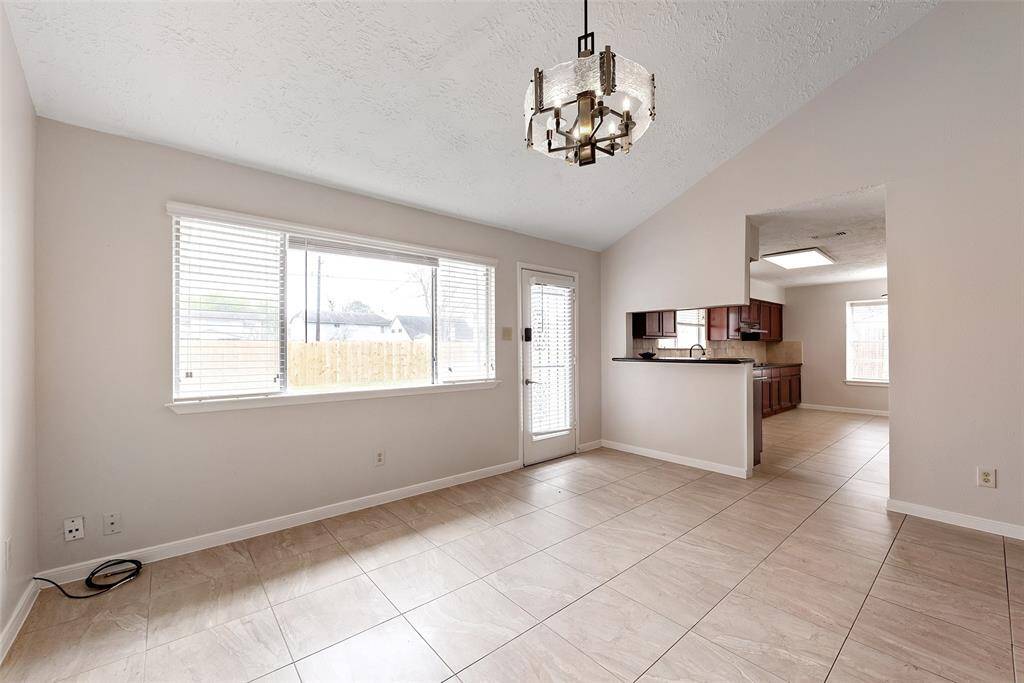 Bright and airy room with large windows and a door leading outside. It features neutral tile flooring, a stylish chandelier, and an open wall space to the kitchen, perfect for easy entertaining.