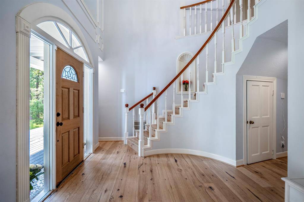 This elegant entryway features a stunning curved staircase with wooden steps and white balusters, leading down to a welcoming front door with a decorative glass panel. The space is accentuated by a large chandelier and soft blue walls with classic molding details.