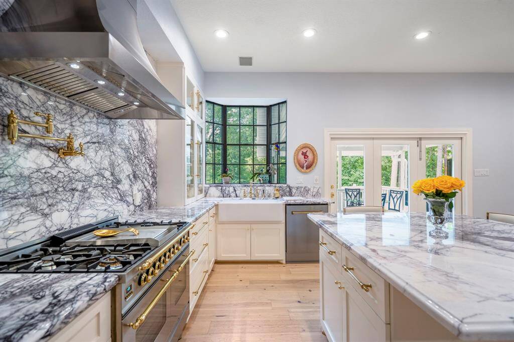 This kitchen features elegant marble countertops, a spacious island, and a high-end Ilve gas range. It offers ample natural light through a large window and French doors, creating a bright and inviting space. Notice the custom pot filler above the large gas stove as well!