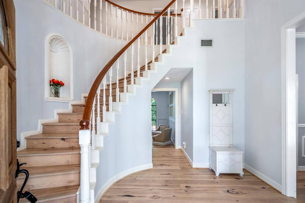 This entryway features a welcoming wooden door with a decorative glass panel, natural light from large windows, and an elegant curved staircase with white spindles and a wooden handrail. The space is bright with light hardwood flooring and soft pastel walls, offering a charming and airy ambiance.