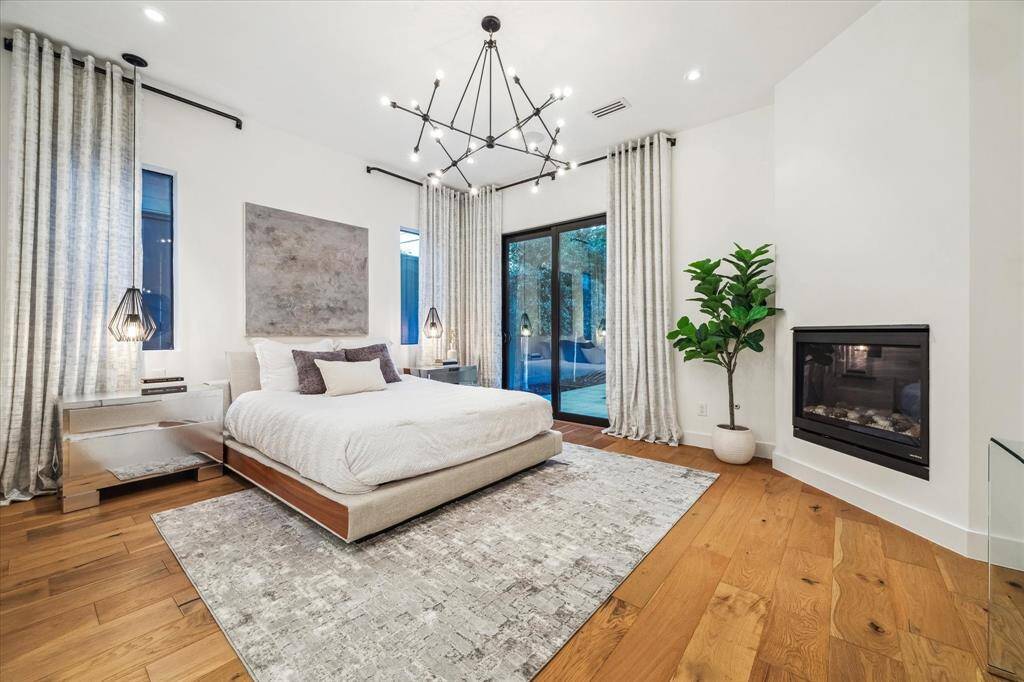This serene first-floor primary bedroom features warm hardwood floors, a cozy modern fireplace, striking statement lighting, and elegant floor-to-ceiling drapes that frame tall windows and sliding glass doors leading to a private backyard retreat.