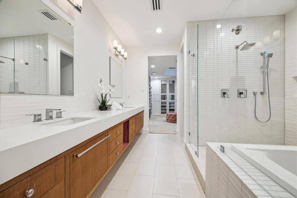 This spa-like primary bathroom exudes tranquility with its soft, neutral color palette, sleek white tile, and warm wood cabinetry that adds a natural touch to the modern double vanity, walk-in shower, and soaking tub.