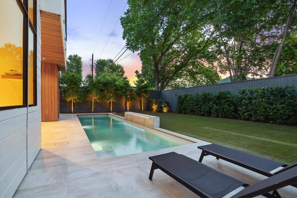 This inviting backyard offers a clean-lined, contemporary pool accented by a tranquil water feature. Framed by rich greenery and tall uplit holly trees, the space feels private and peaceful. Subtle lighting highlights the landscaping at twilight, while the low-maintenance turf provides a vibrant contrast to the light stone patio.