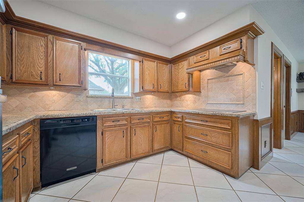 The kitchen (13.4 x 10.8) blends beauty and function. A stainless steel sink sits below a window with a granite ledge and view of the side yard and driveway. To the left, a quiet KitchenAid dishwasher makes cleanup easy. To the right, a sleek 5-burner LG cooktop (2024) with a decorative backsplash sets the stage for signature meals, and easy cleanup. Pots and pans drawers below keep essentials within reach, making time in the kitchen practical and efficient.
