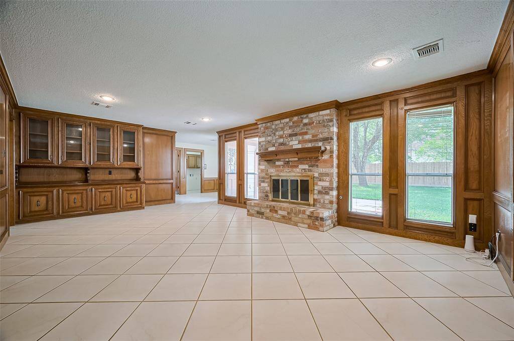 The spacious family room (23.6 x 14.6) offers both comfort and function with easy-to-clean tile flooring and recessed LED lighting. A built-in unit spans one wall—lower cabinets provide ample storage, while upper cabinets with glass doors are perfect for displaying cherished keepsakes. The center shelf is just right for family photos or serving buffet-style during holiday gatherings. A beautiful fireplace anchors the space, flanked by walls of windows that fill the room with natural light and warmth.
