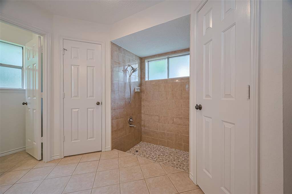 Opposite the double sinks is a step-down walk-in shower with a pebble floor and tiled walls, creating a spa-like atmosphere. A convenient toe-tester lets you adjust the water temperature before stepping in. Generous, lighted walk-in closets flank each side of the shower, providing ample storage space. On the left, the water closet with a commode offers added privacy. Two frosted windows brighten the bath with natural light while maintaining privacy.