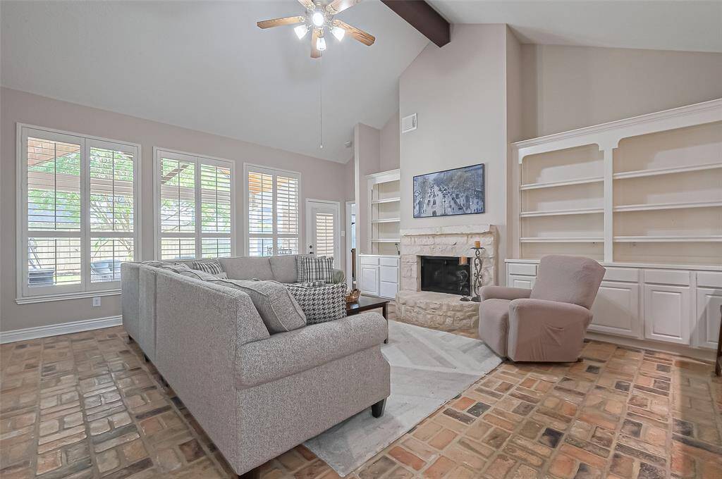 Inviting living room with wall of windows for natural light, brick flooring, limestone gas log fireplace, vaulted ceiling, and open to the kitchen