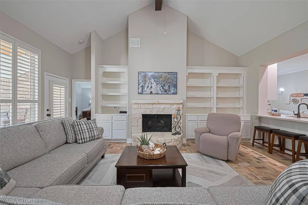 Inviting living room with wall of windows for natural light, brick flooring, limestone gas log fireplace, vaulted ceiling, and open to the kitchen