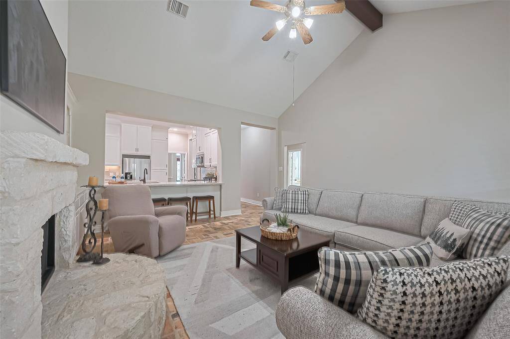 Inviting living room with wall of windows for natural light, brick flooring, limestone gas log fireplace, vaulted ceiling, and open to the kitchen
