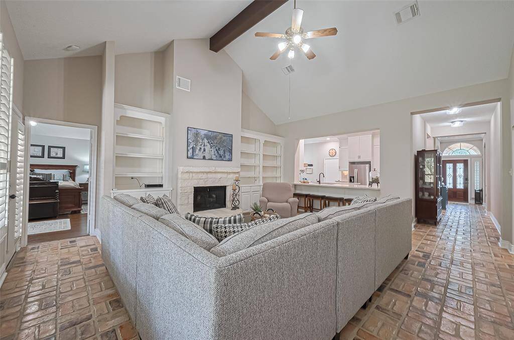 Inviting living room with wall of windows for natural light, brick flooring, limestone gas log fireplace, vaulted ceiling, and open to the kitchen