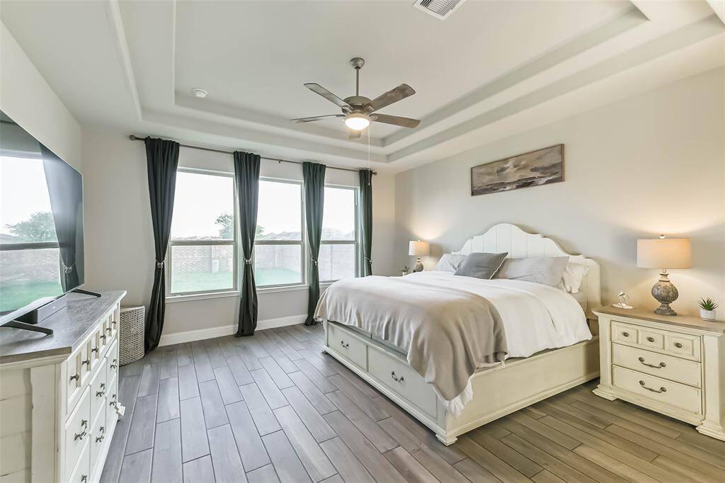 Spacious primary bedroom with wood look tile also includes a trayed ceiling, ceiling fan, and a wall of windows looking to the back yard and brick fence.