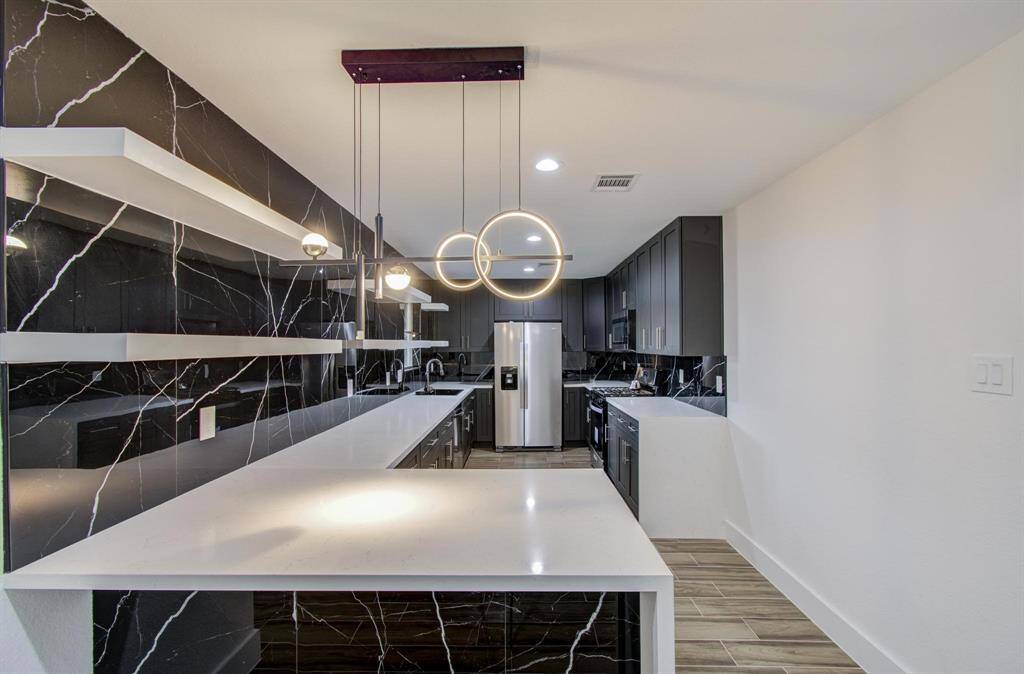 The countertops are white quartz, providing a refreshing contrast to the dark tones of the cabinets and backsplash.  Above the island hangs a unique, contemporary pendant light fixture with circular element