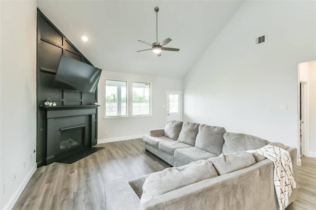 Generous living room boasts an accent wall around the fireplace, high ceiling, and ceiling fan with a view to the huge backyard with the trees behind it.
