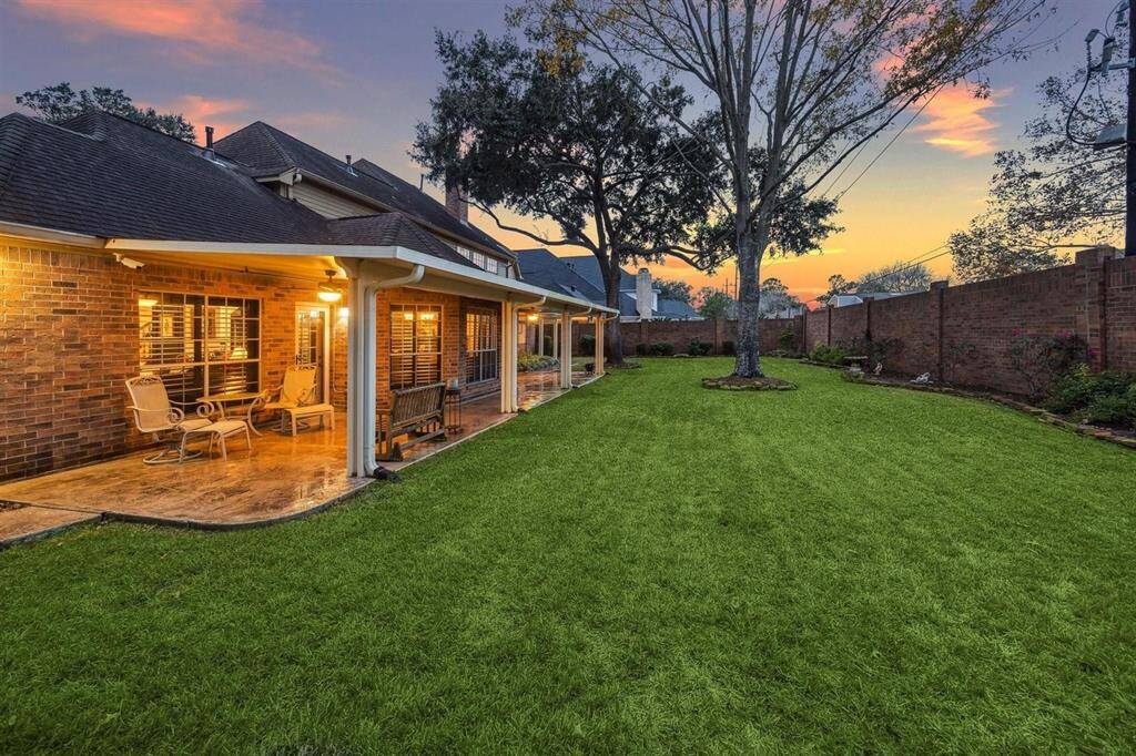 Discover your private backyard oasis at dusk! This beautifully landscaped retreat boasts a generous covered patio with stamped concrete flooring—ideal for relaxing or entertaining year-round. Glowing windows invite you inside while the expansive green lawn, shade trees, and brick fencing provide peace and privacy. The perfect blend of nature, space, and serenity for your outdoor lifestyle.