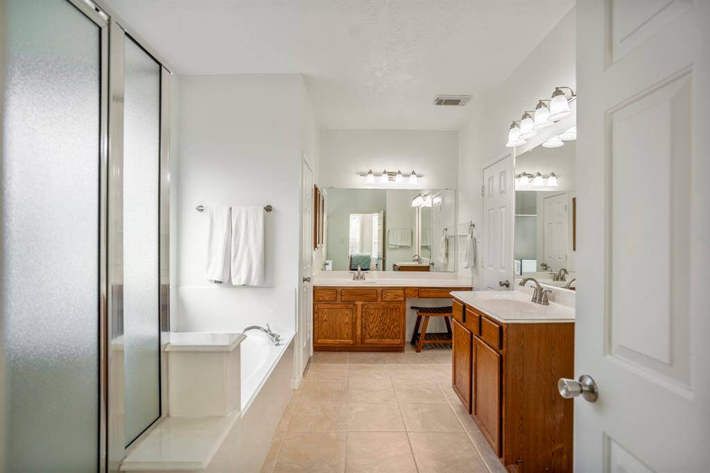 Entering the primary bath, you’re greeted by a spacious layout featuring a separate shower and soaking tub to the left, dual vanities including a dedicated makeup area straight ahead, and private water closet tucked beside. Two doors on the right reveal a generous walk-in closet.