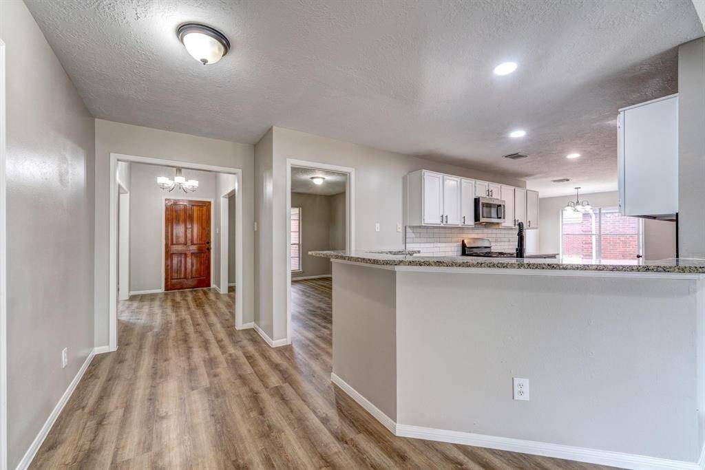 This photo showcases an open-concept living space with modern finishes. The kitchen features white cabinetry, granite countertops, and stainless steel appliances. The room has wood-look flooring and neutral walls, leading to a dining area with natural light.