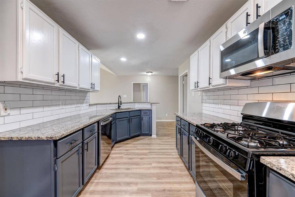 This modern kitchen features sleek granite countertops, a stainless steel gas stove and microwave, white subway tile backsplash, and contrasting two-tone cabinetry. The open layout and wood-look flooring add warmth and functionality.