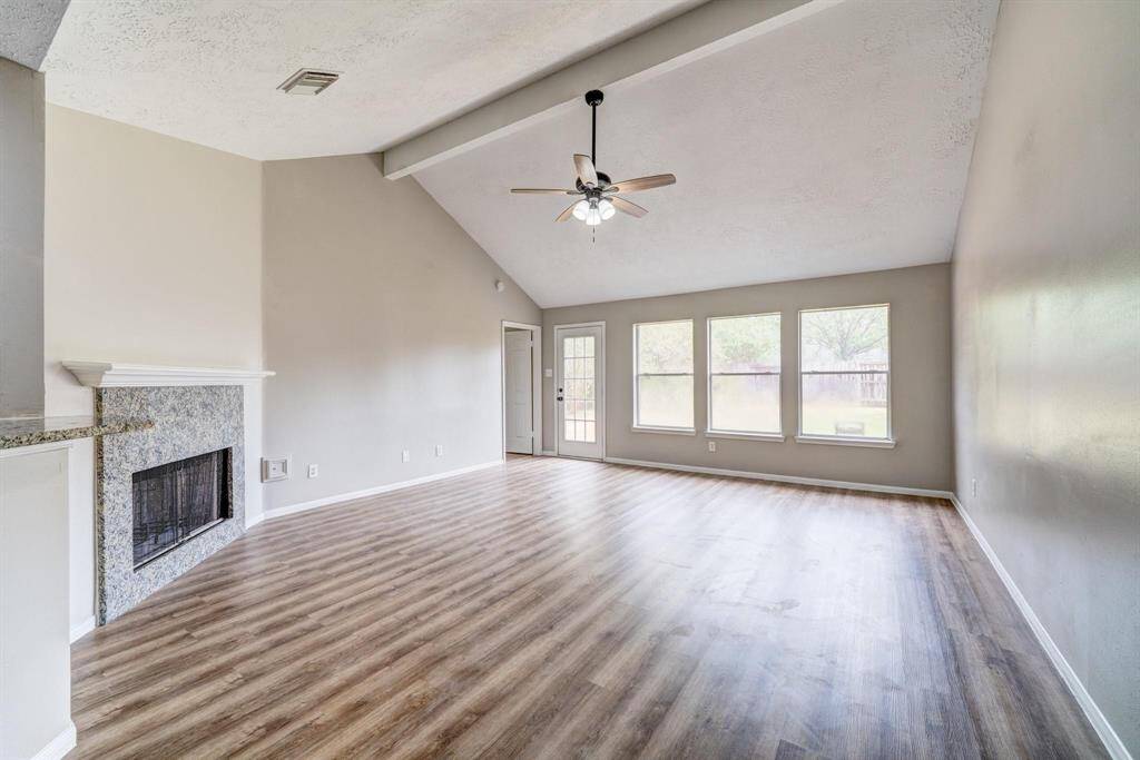 This spacious living room features high vaulted ceilings, a ceiling fan, and abundant natural light from large windows. It includes a cozy fireplace with a modern finish and stylish wood flooring, creating a warm and inviting atmosphere.
