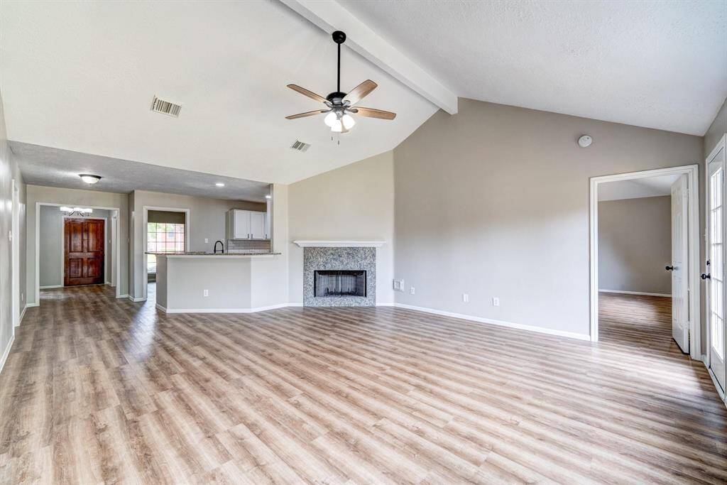 This bright, open-concept living space features vaulted ceilings, wood-style flooring, and a central fireplace. It flows seamlessly into the kitchen area with a breakfast bar. Natural light fills the room through large windows and a door leading to an adjacent room.