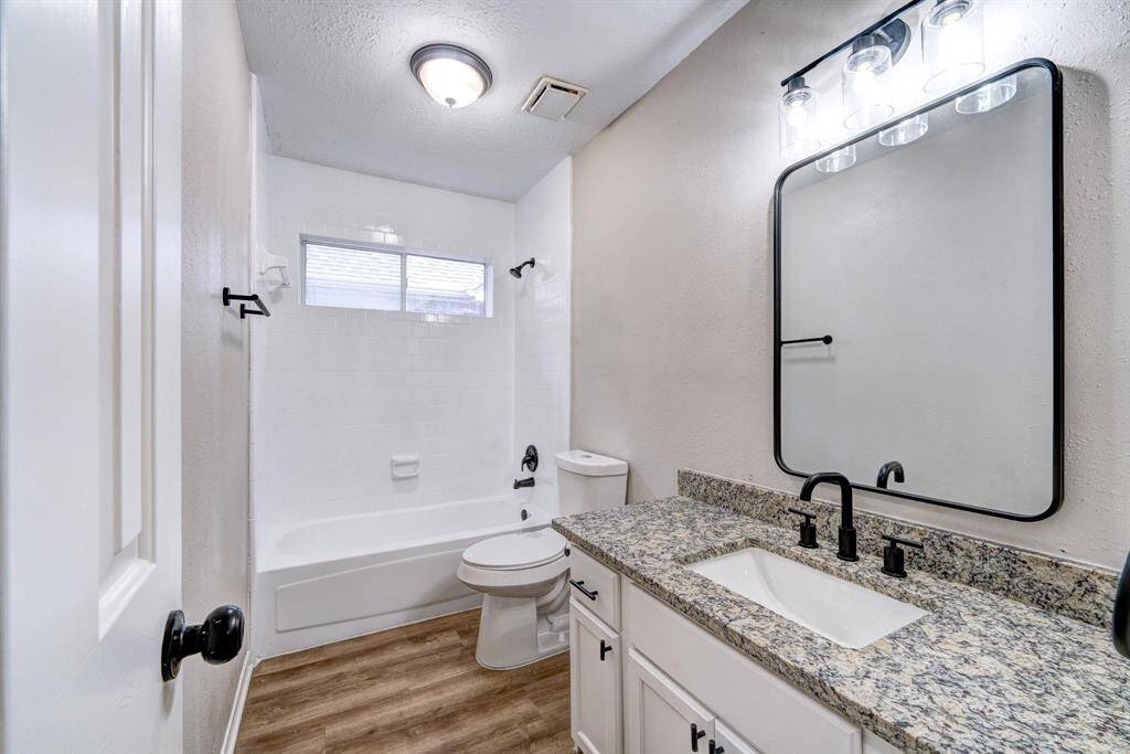 This is a modern bathroom featuring a granite countertop with a stylish black faucet and fixtures. It includes a white vanity with storage, a large framed mirror, and a combined bathtub and shower with white tile. The space is well-lit with both natural light from a window and ceiling lighting.