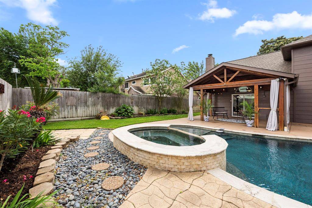 This photo showcases a backyard with a spacious pool and a hot tub, surrounded by a stone and pebble pathway. There's a covered patio with seating, perfect for outdoor relaxation, set against a backdrop of lush greenery and privacy fencing.