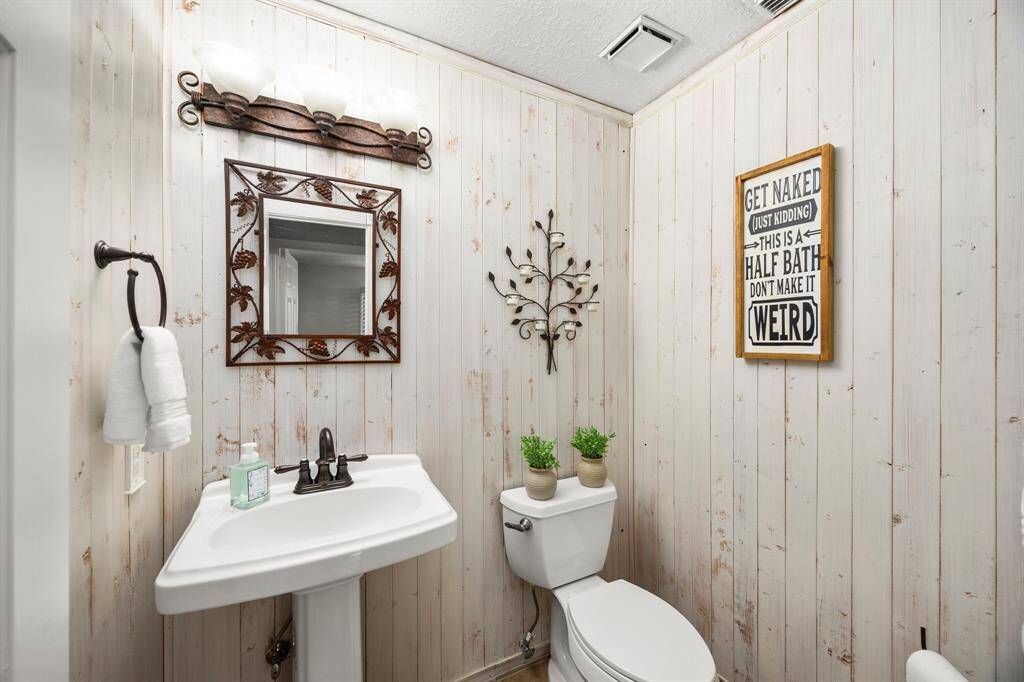 This cozy half-bath features rustic wood-paneled walls, a decorative mirror, and charming iron accents. It includes a pedestal sink and toilet, with light fixtures adding a warm ambiance.