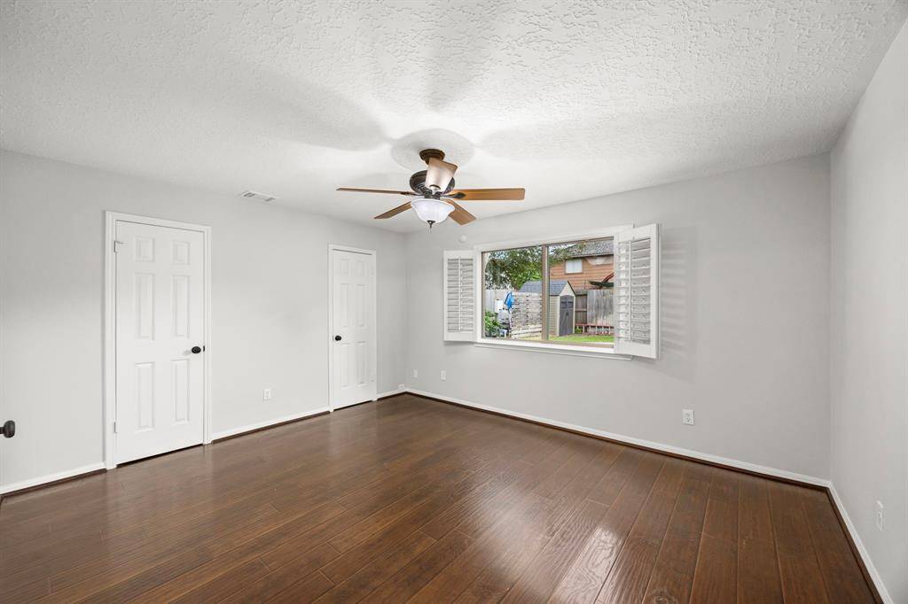 Bright and spacious primary bedroom with rich wood look flooring, a ceiling fan, and large window with shutters offering a view of the backyard. The doors offer access to the wall length closet.